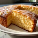 Close-up of a slice cut from a moist healthy greek yogurt cake topped with white lemon glaze and zest.