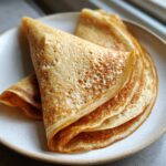 Close-up of three folded high-protein oat & buckwheat crepes stacked on a light ceramic plate.
