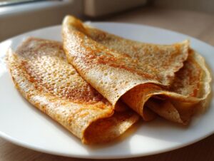 Two folded high-protein oat & buckwheat crepes with golden-brown spots resting on a white plate near a window.