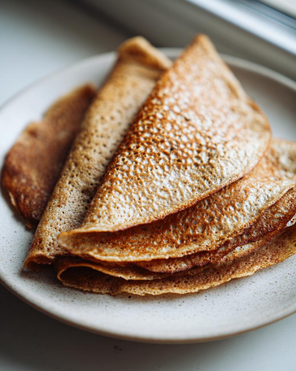 A stack of golden-brown, folded high-protein oat & buckwheat crepes resting on a speckled ceramic plate.