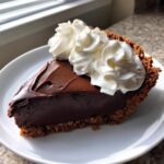 A rich slice of homemade chocolate pie with a dark chocolate filling and a crumb crust, topped with swirls of whipped cream.