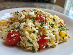 A close-up serving of bright lemon orzo pasta salad mixed with cherry tomatoes, feta cheese, and fresh herbs.