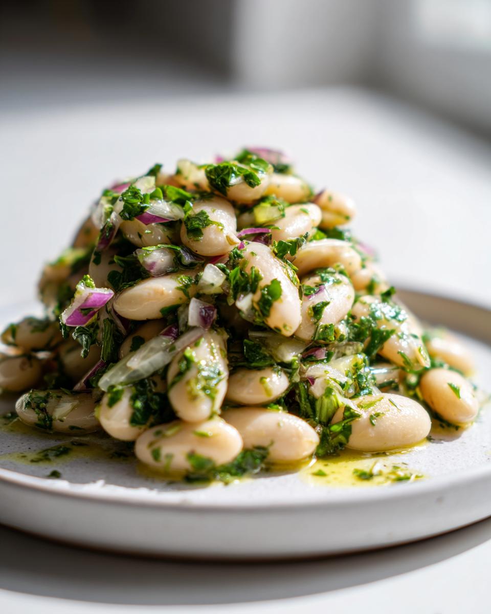 A close-up, bright photo of a mound of marinated white bean salad featuring cannellini beans, red onion, and fresh green herbs.