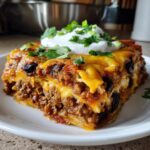 A generous slice of layered mexican lasagna filled with seasoned ground beef, cheese, and black beans, topped with sour cream and cilantro.