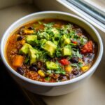 A close-up of a bowl of rich Mexican quinoa soup filled with black beans, corn, and topped generously with diced avocado and cilantro.