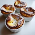 Four golden brown mini dutch baby pancakes dusted with powdered sugar, served in white ramekins.