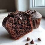 Close-up of a halved, moist chocolate muffin showing the rich interior texture and chocolate chips, with another whole chocolate muffin behind it.