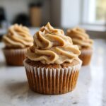 A close-up of one perfectly frosted peanut butter banana cupcake with two others blurred in the background.
