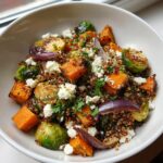 A bowl of roasted vegetable salad featuring sweet potatoes, Brussels sprouts, red onion, quinoa, and crumbled feta cheese.