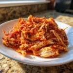 A close-up of shredded crockpot italian chicken mixed with rich red tomato sauce and herbs, served on a white plate.
