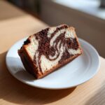 A close-up of a moist slice of german marble cake showing distinct vanilla and chocolate swirls on a white plate.