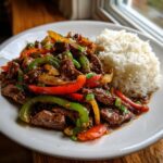 A plate featuring tender slow cooker pepper steak mixed with colorful bell peppers and onions, served alongside fluffy white rice.
