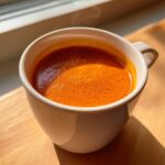 Close-up of a steaming white mug filled with bright orange roasted vegetable soup.
