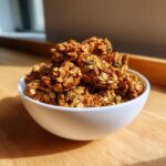 Close-up of crunchy clusters of amazing super seed granola, featuring oats and visible pumpkin seeds, served in a white bowl.
