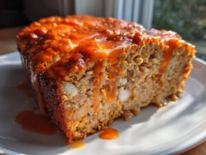 A thick slice of Buffalo chicken meatloaf, generously drizzled with a shiny orange glaze, sitting on a white plate.