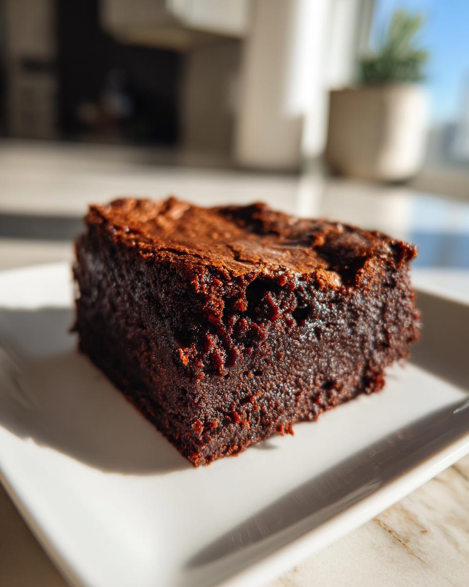 A single, rich, dark square of fudgy sweet potato brownies sitting on a white plate in bright sunlight.