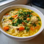 A close-up of a white bowl filled with Mediterranean lemon chicken soup, featuring shredded chicken, orzo pasta, carrots, and fresh parsley.