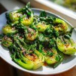 A plate of vibrant green, perfectly Sautéed bok choy halves drizzled with sauce and topped with sesame seeds.