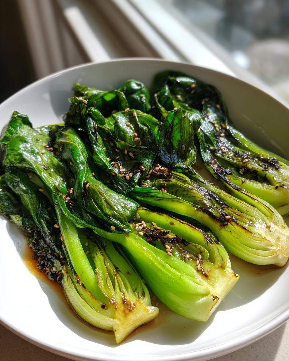 Close-up of bright green, glossy sautéed bok choy halves drizzled with sauce and sprinkled with sesame seeds.