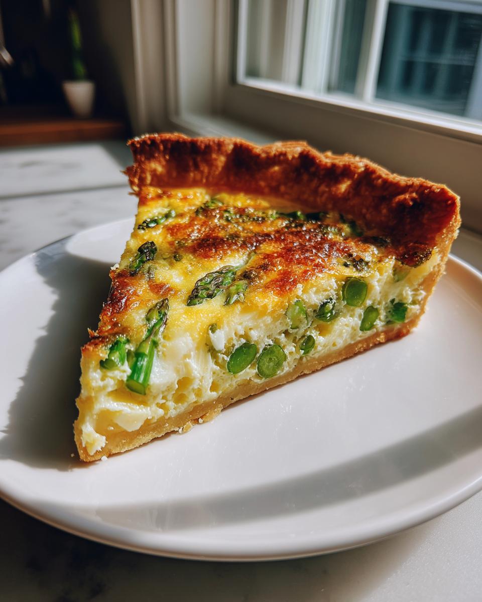 A perfect slice of asparagus quiche with a golden, flaky crust sitting on a white plate near a window.