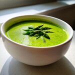 A close-up of a creamy, bright green bowl of Spring asparagus soup, garnished with fresh asparagus tips.
