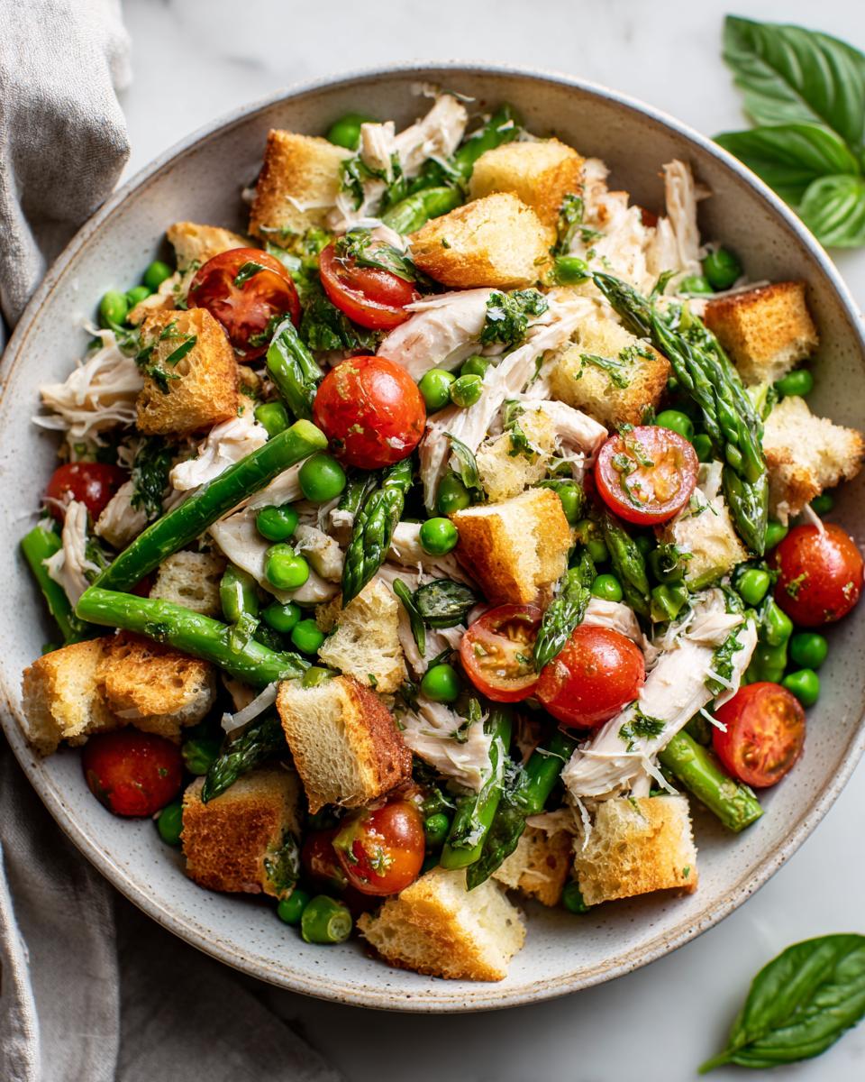 Close-up of a vibrant Spring chicken panzanella salad featuring shredded chicken, croutons, cherry tomatoes, asparagus, and peas.