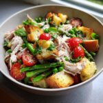 A close-up of a vibrant Spring chicken panzanella salad featuring shredded chicken, croutons, cherry tomatoes, asparagus, and peas.