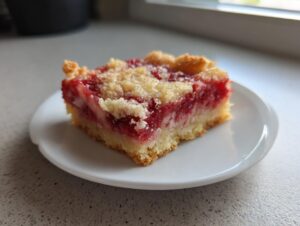 A close-up of a single, square serving of strawberry rhubarb bars with a crumb topping on a white plate.
