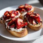 Four pieces of delicious strawberry ricotta bruschetta topped with fresh strawberries and balsamic glaze on a white plate.